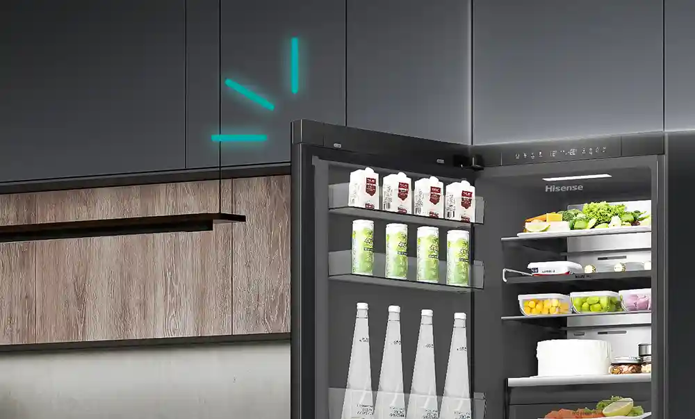 Otwarta lodówka z widocznymi półkami wypełnionymi produktami spożywczymi, takimi jak kartony z napojami, butelki i pojemniki z warzywami. Na drzwiach lodówki widoczne jest logo \'Hisense\'.