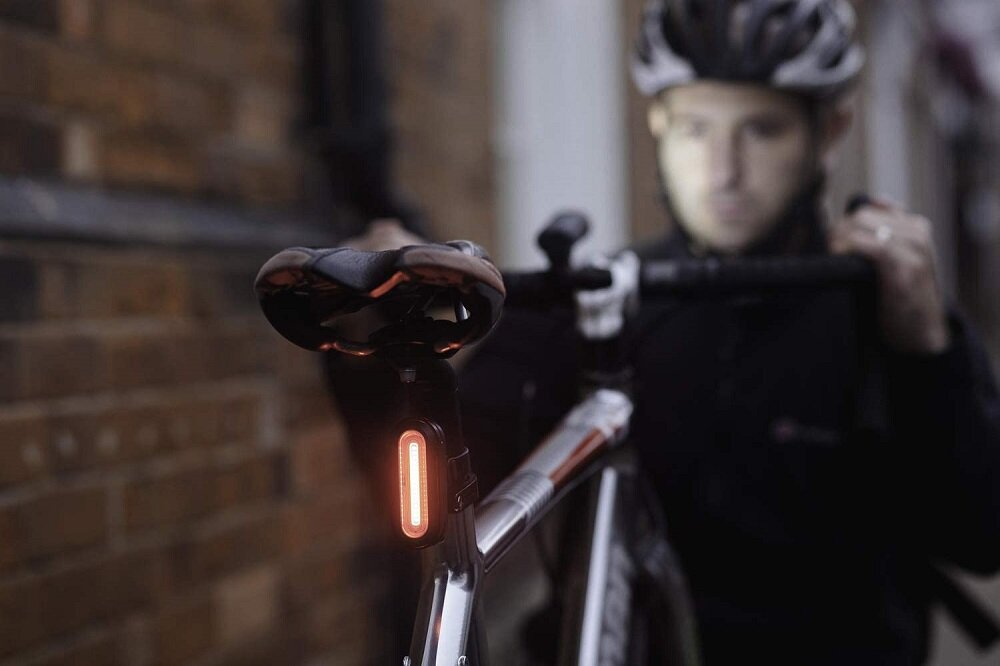Na zdjęciu widać rower z zamontowaną czerwoną lampką pod siodełkiem. W tle znajduje się osoba w kasku rowerowym, trzymająca kierownicę.