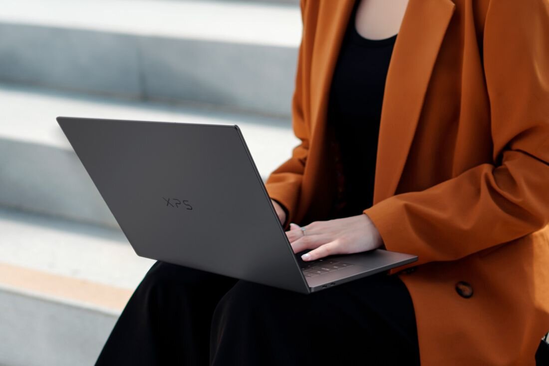 Osoba siedzi na schodach, trzymając otwarty laptop z widocznym logo \'XPS\'. Ubrana jest w pomarańczowy płaszcz.