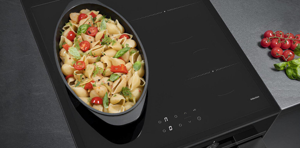 Na obrazie widoczna jest czarna płyta indukcyjna z garnkiem pełnym makaronu, pomidorów i liści bazylii. Obok płyty leżą gałązki pomidorków koktajlowych i kilka liści bazylii.