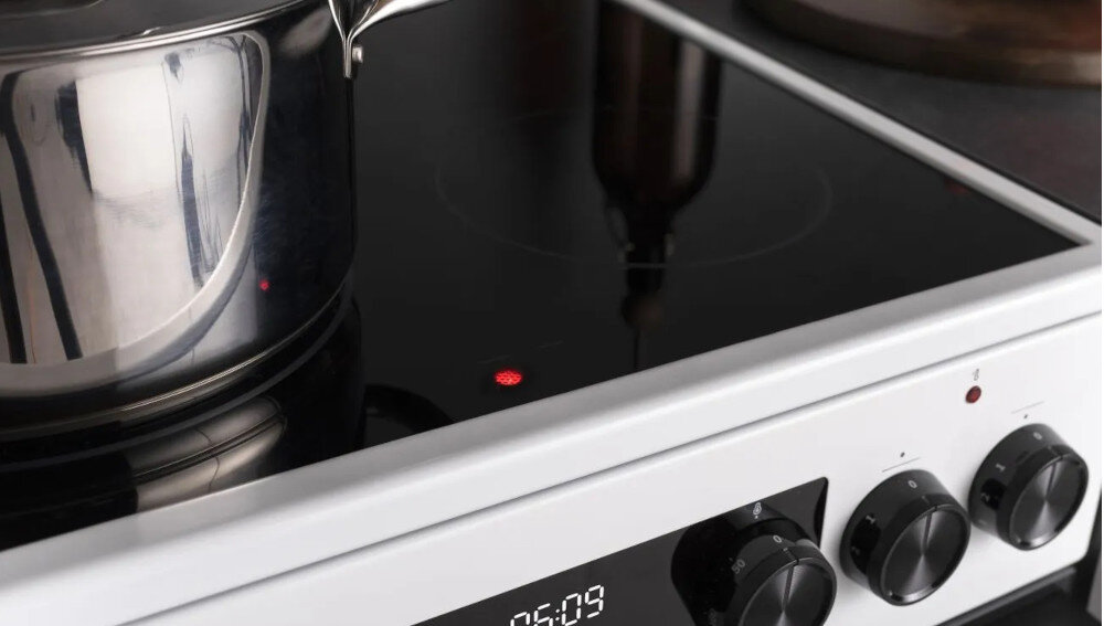 Na zdjęciu widać kuchenkę z płytą indukcyjną, na której stoi metalowy garnek. Panel sterowania pokazuje czas 05:09, a obok znajdują się pokrętła do regulacji ustawień.