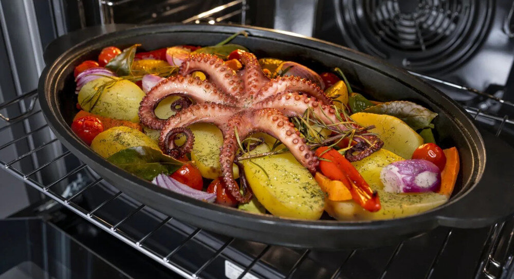 W piekarniku znajduje się owalne naczynie z pieczonymi warzywami i ośmiornicą na wierzchu. Widać ziemniaki, pomidory, cebulę i zioła.