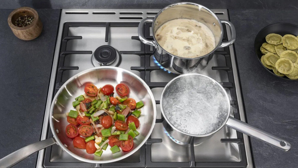 Na kuchence gazowej znajdują się trzy naczynia: patelnia z pokrojonymi pomidorami i papryką, garnek z gotującą się wodą oraz garnek z zupą. Obok na blacie stoi miska z pierożkami.