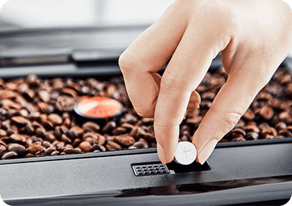 Ręka trzymająca biały element nad tacką z ziarnami kawy. W tle widoczny jest fragment ekspresu do kawy.