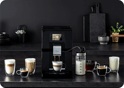 Na blacie kuchennym stoi czarny ekspres do kawy z wyświetlaczem, obok którego znajduje się kilka filiżanek z różnymi rodzajami kawy oraz dzbanek z mlekiem. W tle widoczne są akcesoria kuchenne i rośliny.