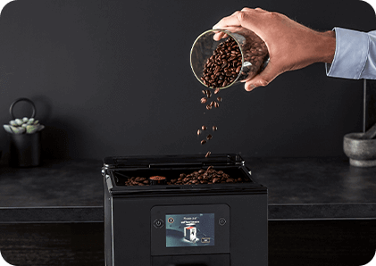 Osoba wsypuje ziarna kawy do pojemnika ekspresu do kawy. Na wyświetlaczu urządzenia widoczny jest obrazek filiżanki kawy.