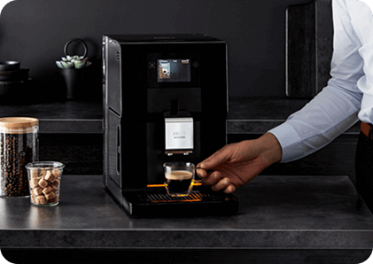 Osoba przygotowuje kawę za pomocą czarnego ekspresu do kawy, na blacie obok znajdują się słoiki z kawą i kostkami cukru. Na ekspresie widoczny jest napis \'KRUPS\'.