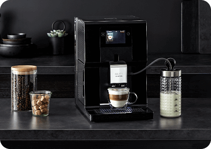 Na blacie kuchennym stoi czarny ekspres do kawy z wyświetlaczem, obok którego znajduje się szklanka z mlekiem oraz słoiki z kawą i cukrem. Na filiżance pod wylewką widnieje napis \'KRUPS Intuition\'.