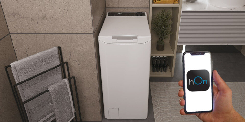 Na obrazie widoczna jest biała pralka w łazience, obok której znajduje się ręcznik na stojaku. Osoba trzyma smartfon z aplikacją \'hOn\' na ekranie.