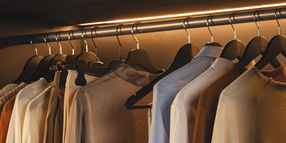 Na obrazie widoczny jest rząd ubrań wiszących na wieszakach w szafie, oświetlony ciepłym światłem LED. Ubrania mają różne kolory, w tym odcienie beżu, brązu i bieli.