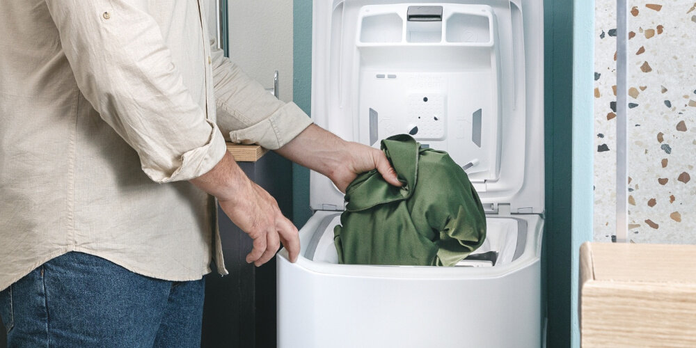 Osoba wkłada zieloną tkaninę do otwartej pralki ładowanej od góry. Wnętrze pralki jest białe i widoczne są elementy konstrukcyjne pokrywy.