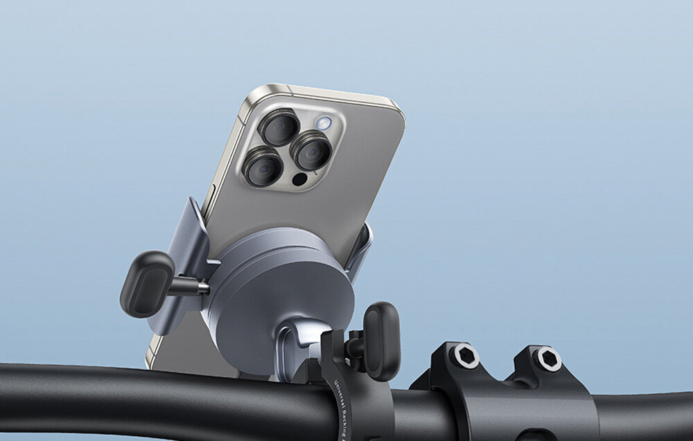 Uchwyt na telefon zamontowany na kierownicy roweru, trzymający smartfon z trzema widocznymi obiektywami aparatu. Uchwyt jest w kolorze szaro-czarnym, a tło jest jednolite, jasnoniebieskie.