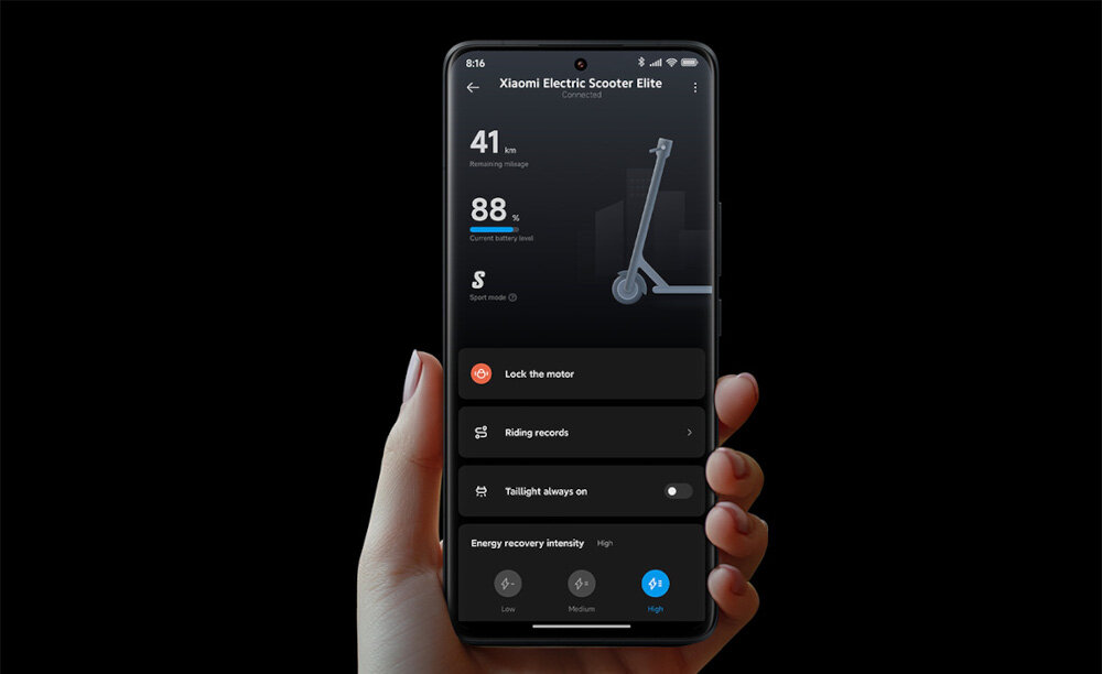 Osoba trzyma smartfon z aplikacją pokazującą dane hulajnogi elektrycznej Xiaomi Electric Scooter Elite. Na ekranie widoczne są informacje o napięciu, prędkości oraz opcje takie jak \'Lock the motor\' i \'Riding records\'.
