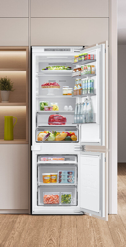 Otwarta lodówka z widocznymi półkami wypełnionymi różnorodnymi produktami spożywczymi, takimi jak warzywa, napoje, jajka i mięso. W dolnej części znajdują się przezroczyste szuflady z mrożonkami.