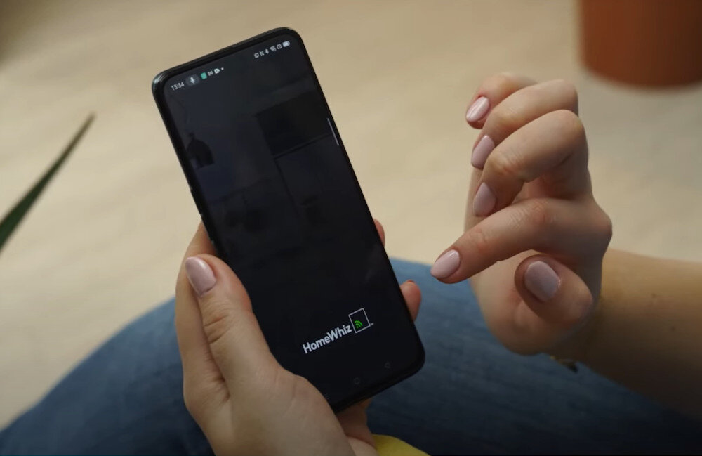 Osoba trzyma smartfon z włączoną aplikacją HomeWhiz. Na ekranie widoczny jest napis \'HomeWhiz\'.