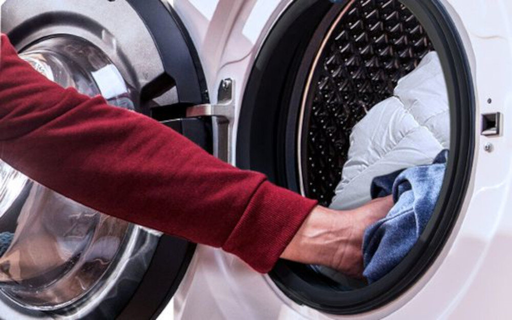 Osoba wkłada ubrania do otwartego bębna pralko-suszarki. Widać metalowe wnętrze urządzenia oraz ubrania w jasnych kolorach.