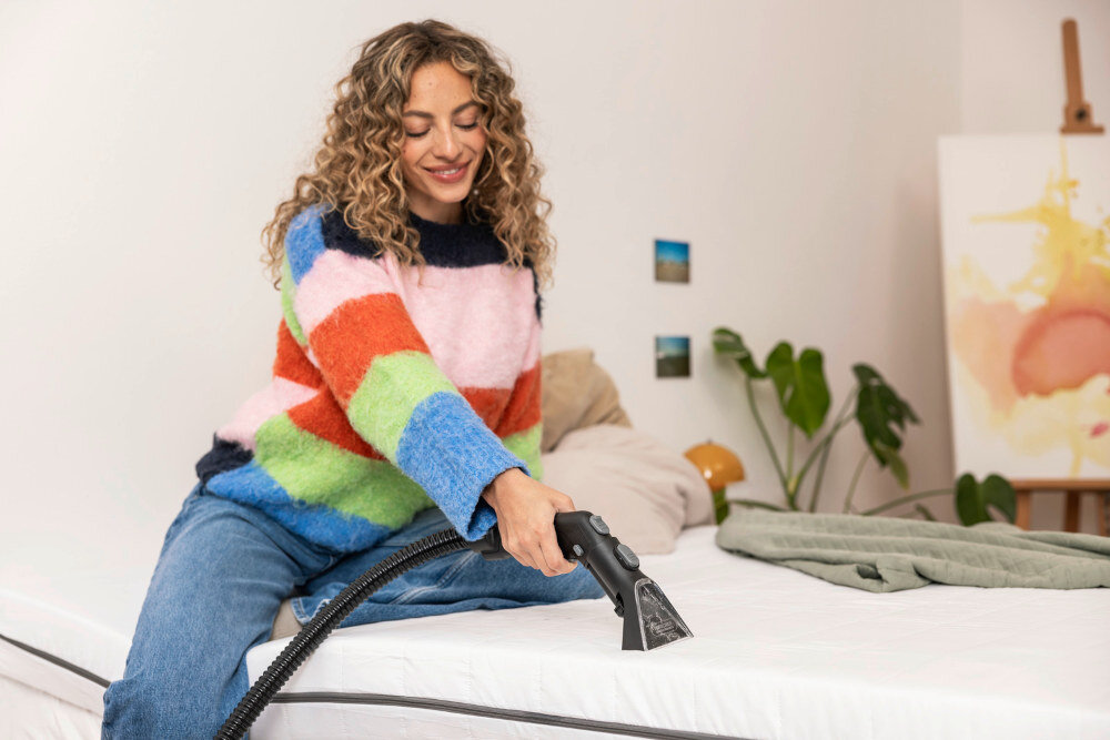 Osoba używa odkurzacza piorącego na białym materacu. W tle widoczne są rośliny i obraz na sztaludze.