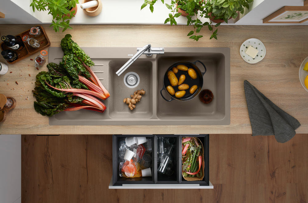 Na blacie kuchennym znajduje się zlewozmywak jednokomorowy z ociekaczem, w którym leżą warzywa i garnek z ziemniakami. Obok zlewu widoczne są świeże warzywa, przyprawy oraz rośliny doniczkowe.