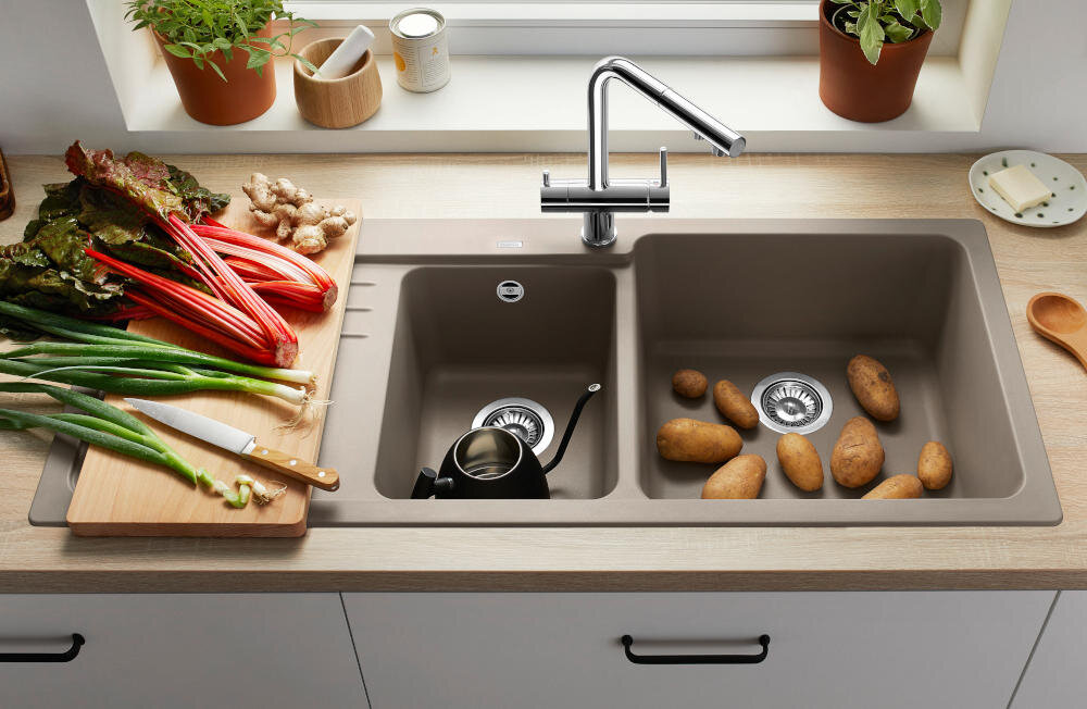 Na blacie kuchennym znajduje się zlewozmywak dwukomorowy z ociekaczem, w jednej komorze leżą ziemniaki, a w drugiej czajnik. Obok zlewu na desce do krojenia znajdują się warzywa i nóż, a w tle widać doniczki z roślinami.