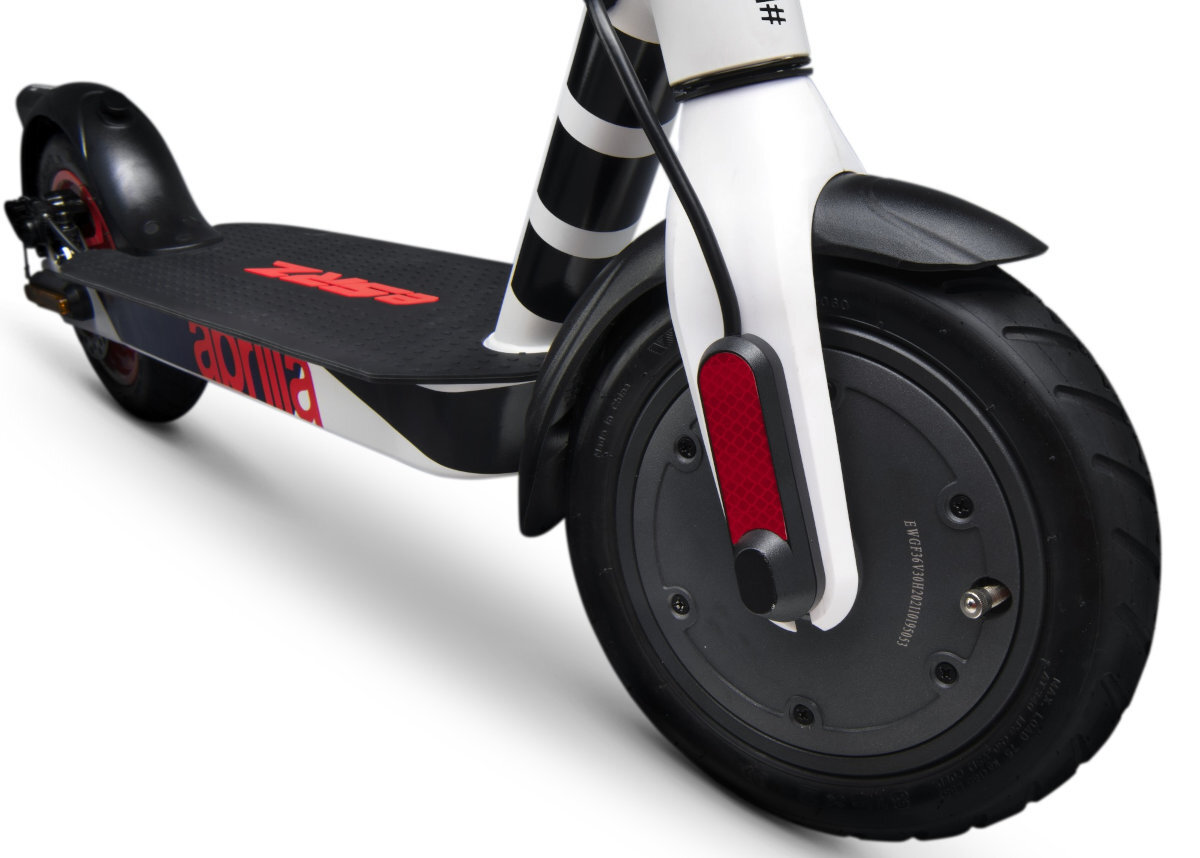 Zbliżenie na przednie koło hulajnogi elektrycznej w kolorach biało-czarno-czerwonym z widocznym logo \'aprilia\' na podeście. Opona jest czarna, a na widelcu znajduje się czerwony odblask.