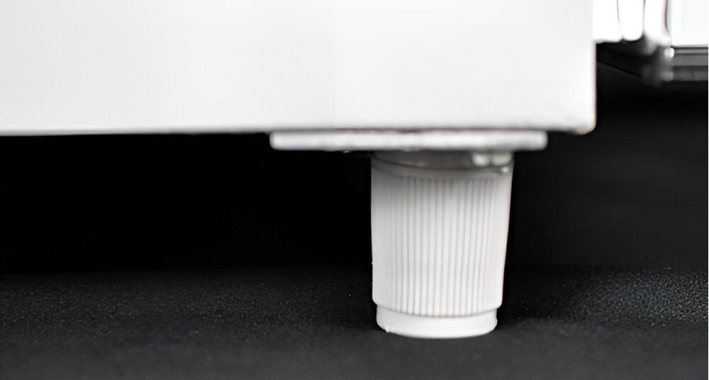 Zdjęcie przedstawia biały, plastikowy element regulacyjny zamontowany na dolnej części urządzenia, prawdopodobnie nóżkę. Tło jest czarne, co kontrastuje z jasnym kolorem elementu.
