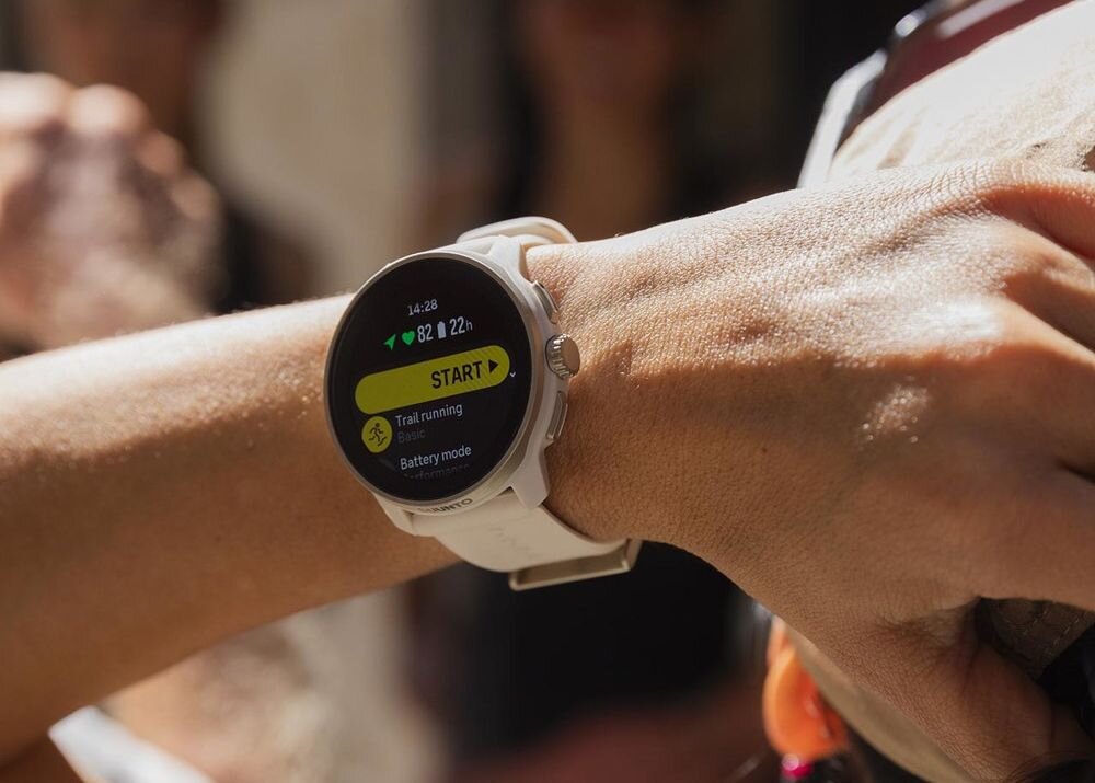 Osoba nosi smartwatch z białym paskiem, na ekranie widoczny jest tekst: \'14:28\', \'82 22h\', \'START\', \'Trail running\', \'Battery mode\'.