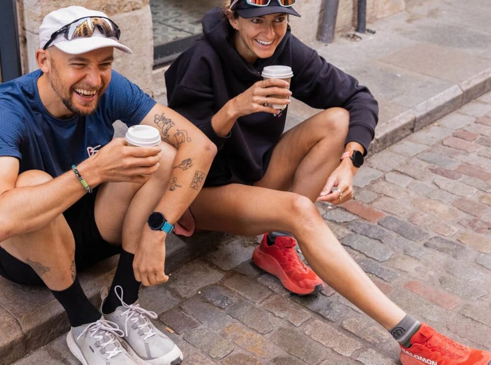 Dwie osoby siedzą na chodniku, trzymając kubki z napojami. Jedna z nich ma na nadgarstku różowy smartwatch.