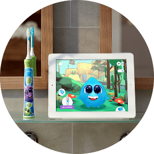 Na szklanej półce stoi kolorowa szczoteczka soniczna z dziecięcym motywem obok tabletu, na którym wyświetlana jest gra z niebieskim stworkiem na tle lasu.