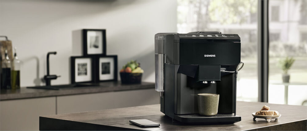 Na blacie kuchennym stoi czarny ekspres do kawy z logo \'SIEMENS\', obok filiżanka i talerzyk z rogalikami. W tle widać zlew, ramki ze zdjęciami i okno z widokiem na zieleń.