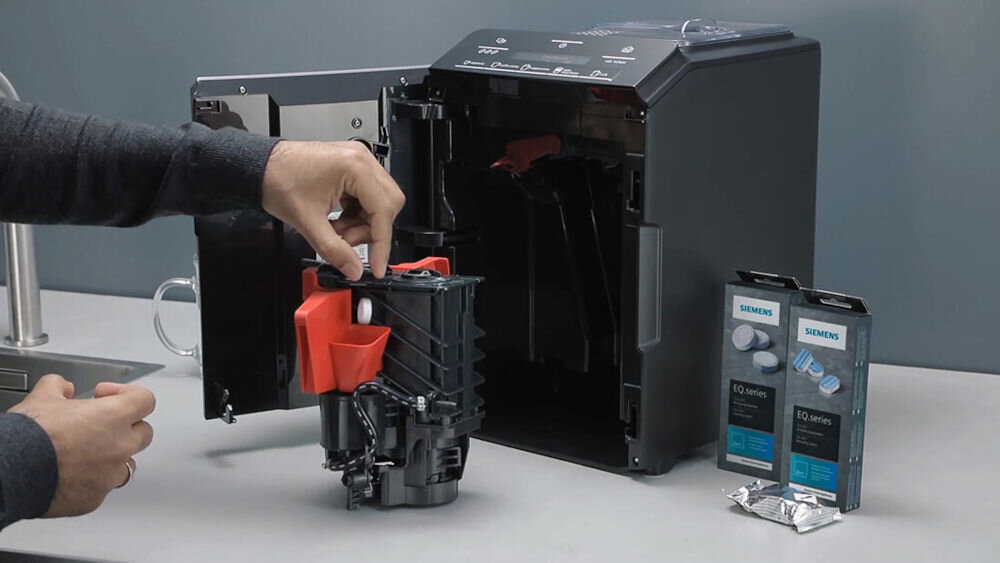 Osoba trzyma wyjęty element ekspresu do kawy, obok otwartego urządzenia. Na blacie stoją dwa opakowania z napisem \'SIEMENS EQ. series\'.