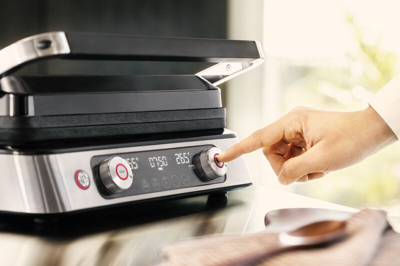 Osoba naciska przycisk na panelu sterowania grilla z wyświetlaczem cyfrowym pokazującym temperaturę 265°C. Grill ma metaliczną obudowę i otwartą pokrywę.