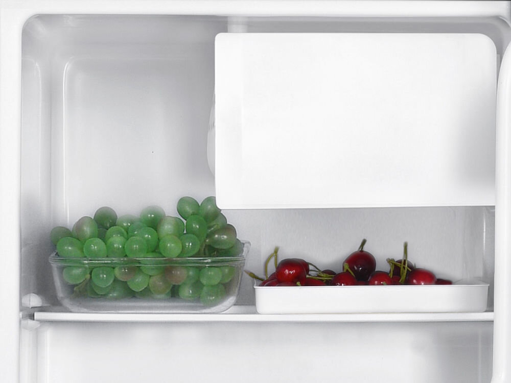 Lodówka GOTZE&JENSEN RB150K 51.5 cm Zbliżenie na górną półkę lodówki z pojemnikami pełnymi zielonych winogron i czerwonych czereśni pod komorą niskiej temperatury. 