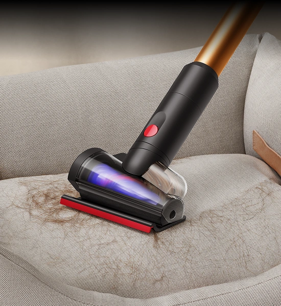 Odkurzacz z czarną i złotą rurą oraz przezroczystą głowicą czyszczącą usuwa zabrudzenia z jasnej tapicerki. Głowica ma czerwony przycisk i niebieskie światło wewnątrz.