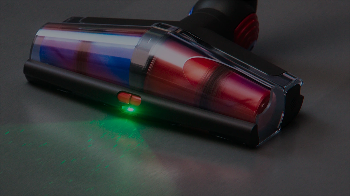 Na obrazie widoczna jest głowica odkurzacza z przezroczystą obudową, w której znajdują się kolorowe elementy. Na obudowie świeci zielona dioda.