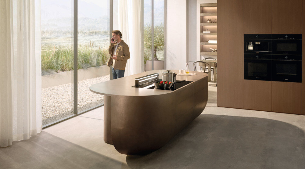 PŁYTA MIELE KM 8565 FL MATTFINISH SERIA KM8000 Kuchnia z dużą wyspą i płytą indukcyjną; przy panoramicznym oknie stoi osoba rozmawiająca przez telefon. W zabudowie widoczne są piekarniki, a całość utrzymana jest w ciepłej, minimalistycznej stylistyce