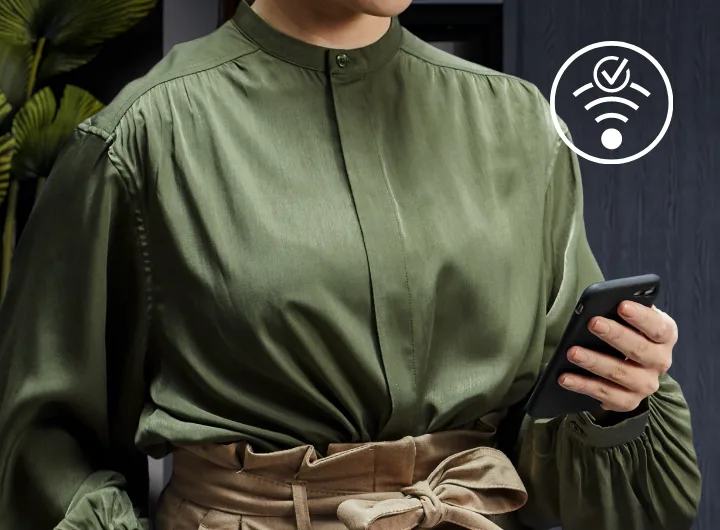 Osoba w zielonej koszuli trzyma telefon, a obok widoczna jest ikona z symbolem Wi-Fi i zaznaczeniem. W tle znajdują się rośliny i ciemna ściana.