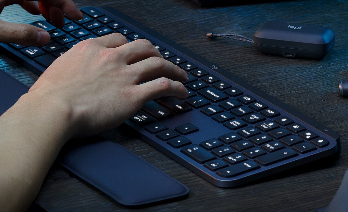 Osoba pisze na grafitowej klawiaturze z podświetlanymi klawiszami, obok znajduje się etui z logo \'logi\'.