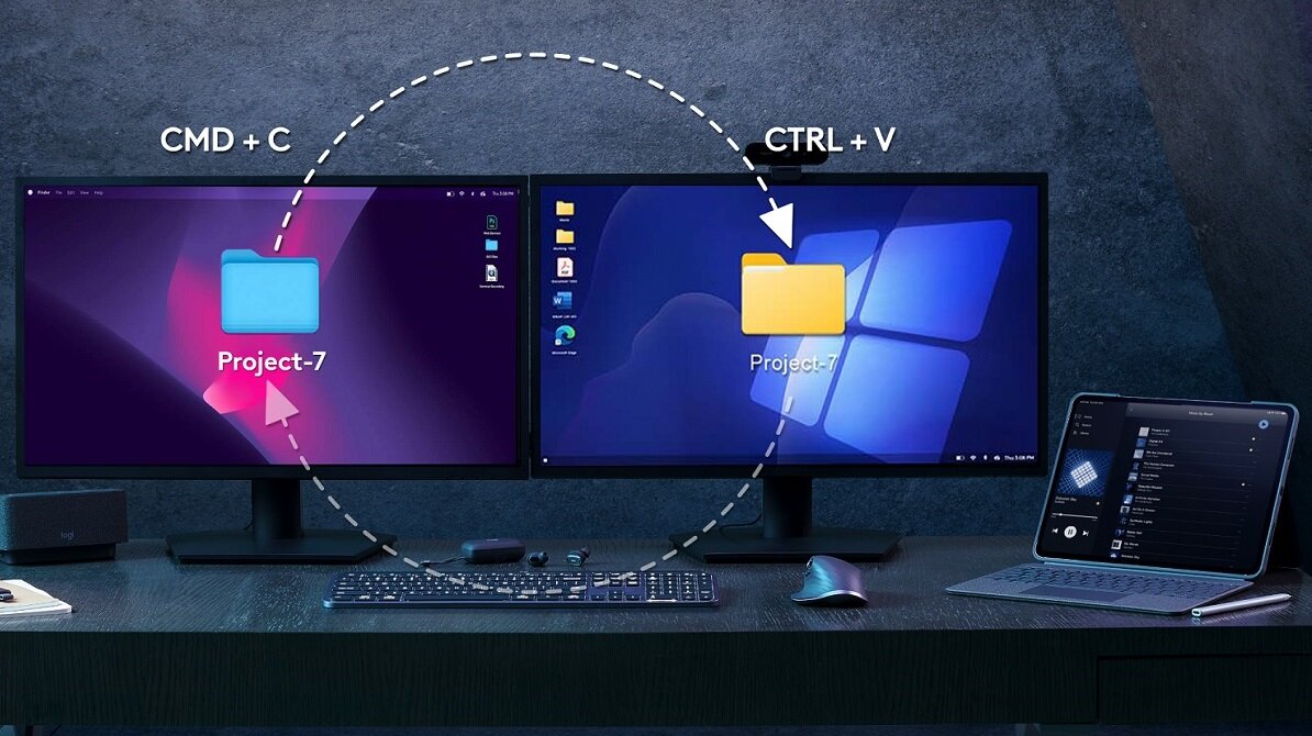 Na biurku znajdują się dwa monitory, klawiatura, mysz i laptop. Na ekranach widoczny jest proces kopiowania folderu \'Project-7\' z użyciem skrótów CMD + C i CTRL + V.