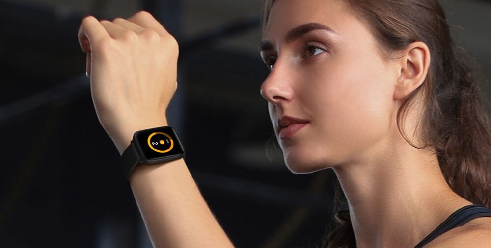 Osoba nosi smartwatch z czarnym paskiem i wyświetlaczem pokazującym żółty okrąg z cyfrą 10 w środku. Tło jest rozmyte, co sugeruje, że zdjęcie skupia się na zegarku.