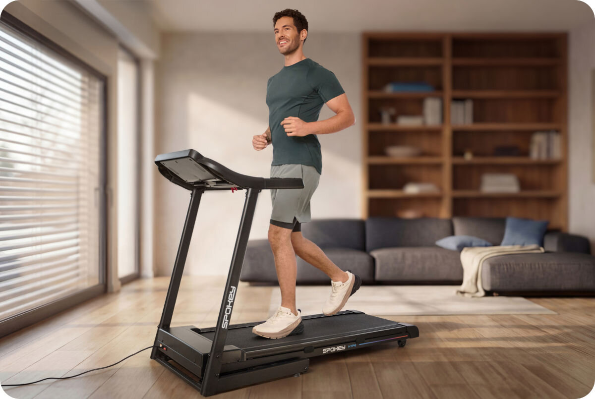 Mężczyzna biegnie na bieżni elektrycznej SPOKEY Hype 1305W 14 km/h w jasnym pokoju z dużym oknem i sofą w tle. Na bieżni widoczne jest logo \'SPOKEY\'.