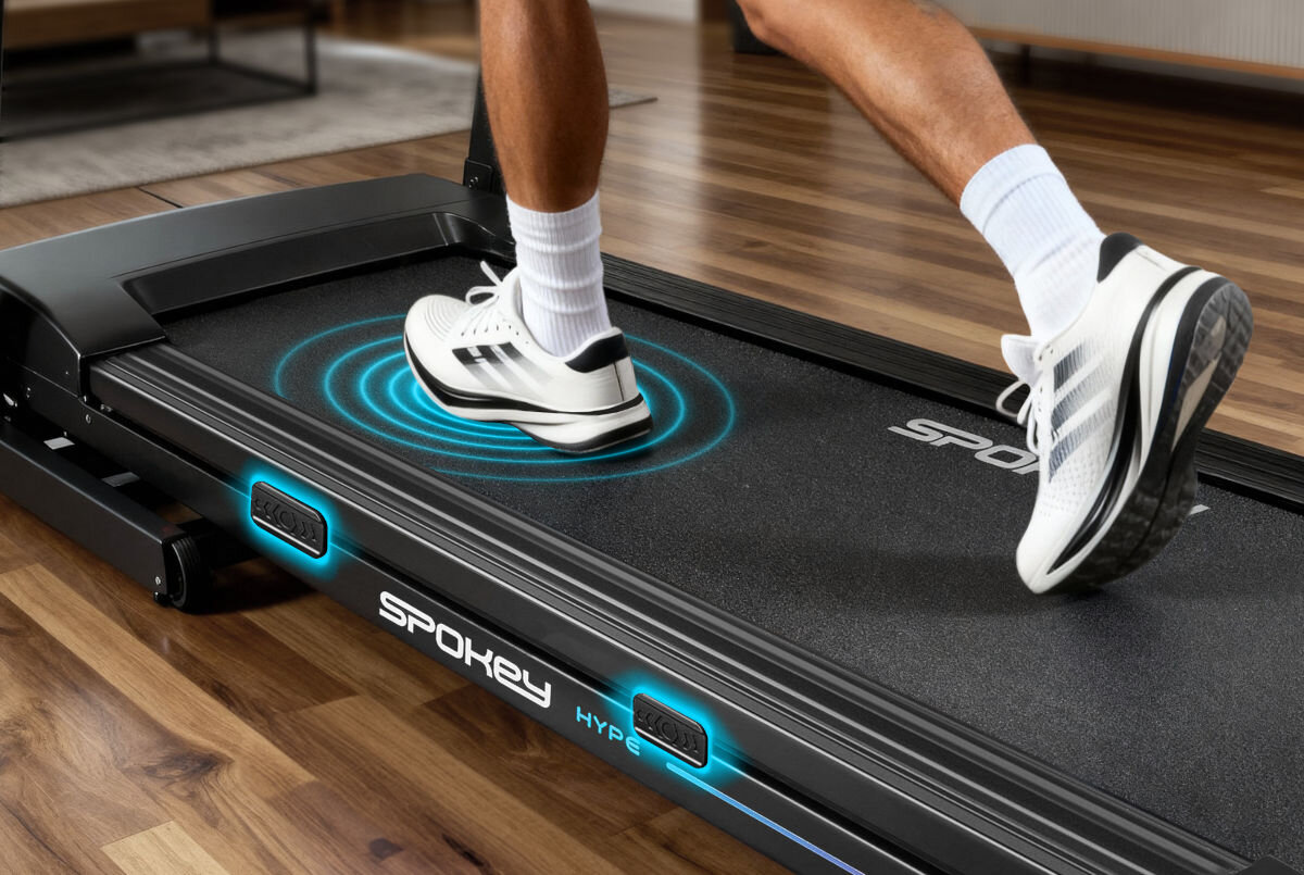 Osoba biegnie na bieżni elektrycznej z widocznym logo \'SPOKEY\' i napisem \'HYPE\'. Pod stopą widoczny jest niebieski efekt graficzny symbolizujący ruch.