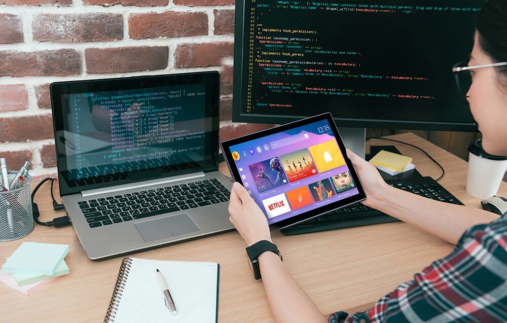 Monitor UPERFECT UPI B7 Osoba siedząca przy biurku trzyma w rękach ekran dotykowy wyświetlający kolorowe ikony aplikacji, obok otwarty laptop z kodem i monitor z edytorem tekstu, opis