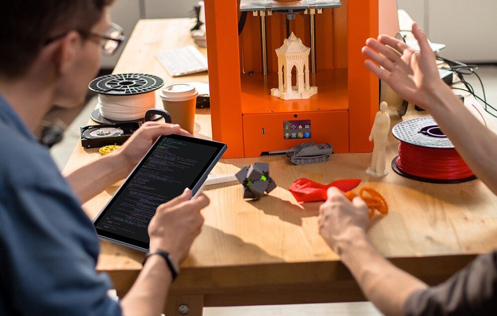 Monitor UPERFECT UPI B7 Dwie osoby przy stole z drukarką 3D, jedna trzyma ekran dotykowy z wyświetlonym kodem, na stole znajdują się wydruki i szpule filamentu, przekątna, matryca IPS, rozdzielczość, jasność