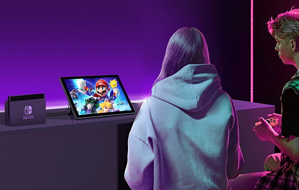 Monitor UPERFECT UPI B7 Dwie osoby siedzące przy biurku grają na konsoli, ekran ustawiony na stole wyświetla kolorową grę, w tle fioletowe światło, głośniki, dźwięk