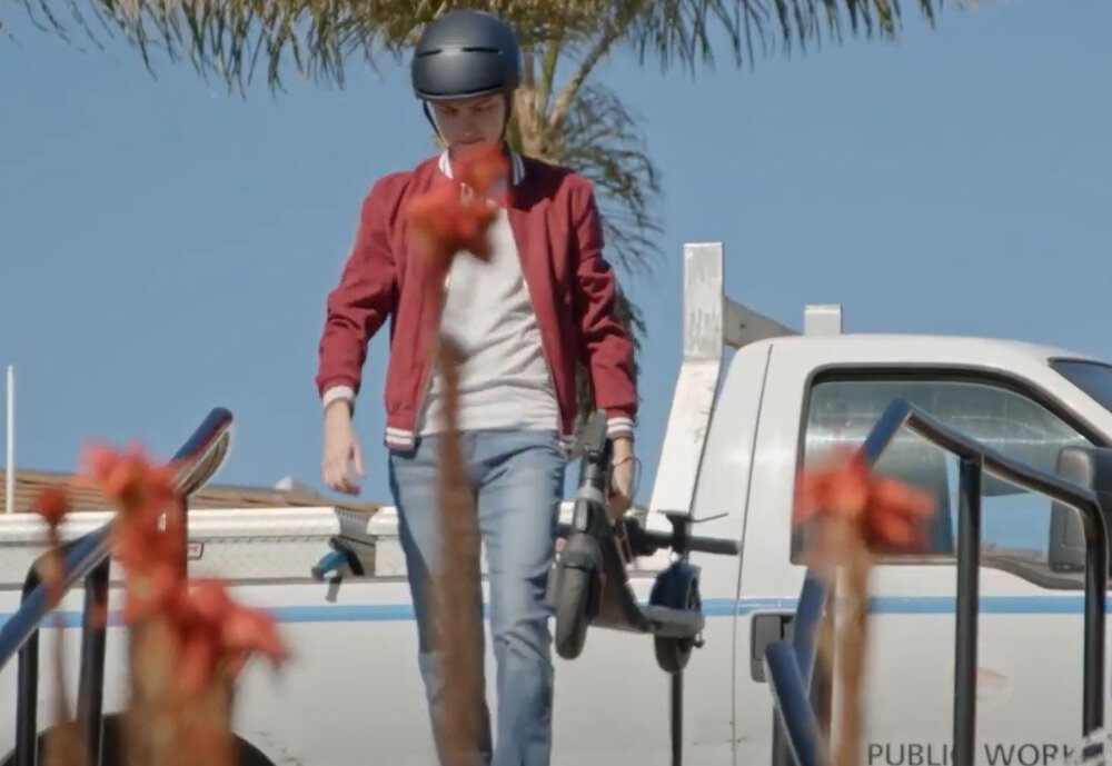 Osoba w kasku trzyma hulajnogę elektryczną, idąc w pobliżu białego samochodu dostawczego. W tle widoczne są palmy i niebo.