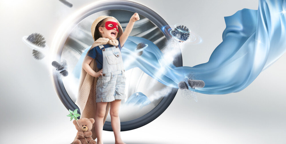 Dziecko w czerwonej masce i pelerynie stoi przed dużym bębnem pralki, unosząc rękę w geście triumfu. Obok znajduje się pluszowy miś z zieloną rośliną.