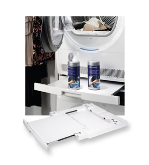 Na obrazie widoczna jest suszarka z otwartymi drzwiczkami, na której wysuniętej półce znajdują się dwa pojemniki z niebieskimi etykietami. Obok suszarki leży biały element, prawdopodobnie część wyposażenia lub akcesorium.