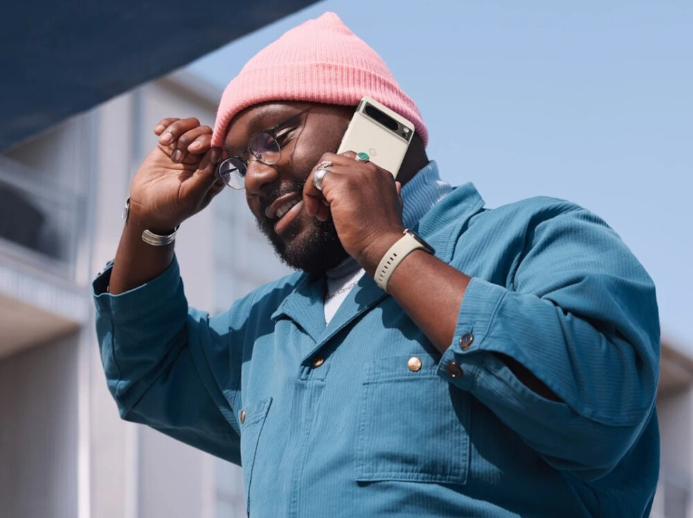 Osoba trzymająca smartfon przy uchu, ubrana w niebieską kurtkę i różową czapkę. W tle widoczny jest fragment budynku i niebo.