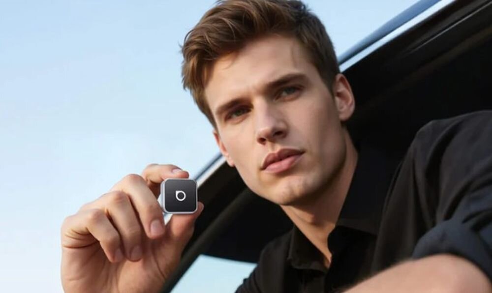 Osoba trzyma małe, czarne urządzenie z białym symbolem na środku. W tle widoczny jest fragment samochodu.