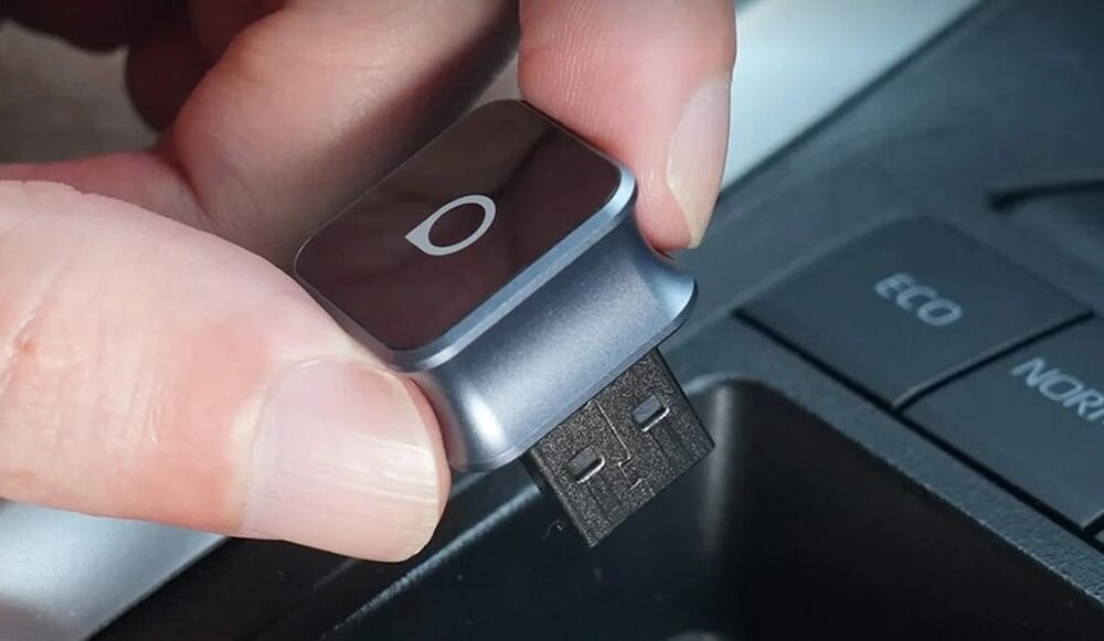 Osoba trzyma małe, czarne urządzenie z logo w kształcie litery \'O\', które jest podłączane do portu USB w samochodzie. Obok widoczne są przyciski z napisami \'ECO\' i \'NORM\'.